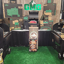 Green Mountain Grills display featuring various grill models, branded packaging, and grilling accessories in a booth setting.