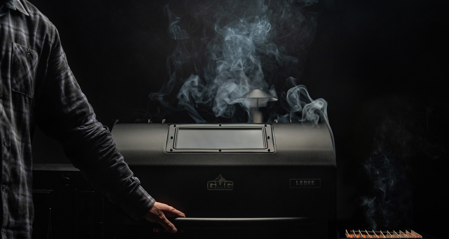 Man in plaid shirt operating a Green Mountain Grills Ledge pellet grill, with smoke rising in a dark setting, emphasizing grilling experience and BBQ culture.