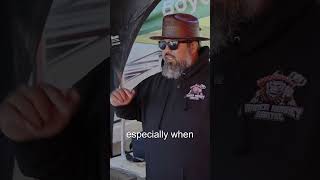 Man gesturing while wearing sunglasses and a hat, discussing grilling techniques at a BBQ event, related to Green Mountain Grills and pellet grilling.