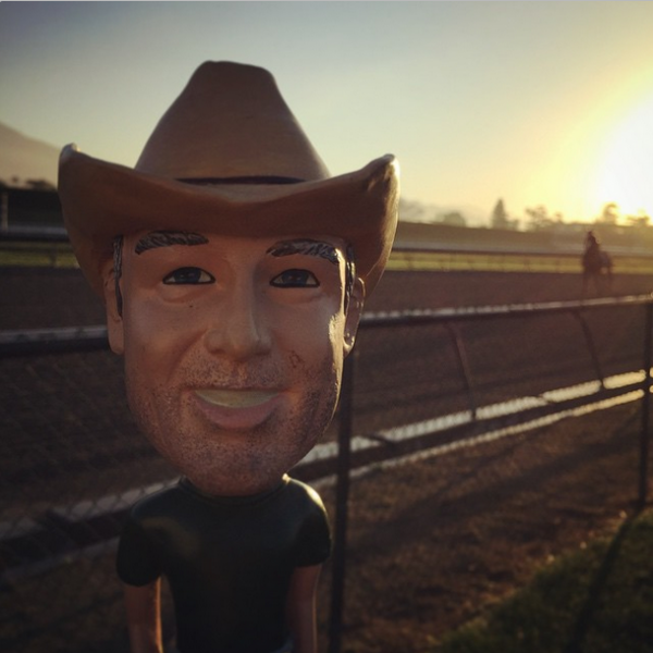 Bobblehead figure of a cowboy with a hat, set against a racetrack background during sunset, representing casual outdoor themes related to grilling and accessories.